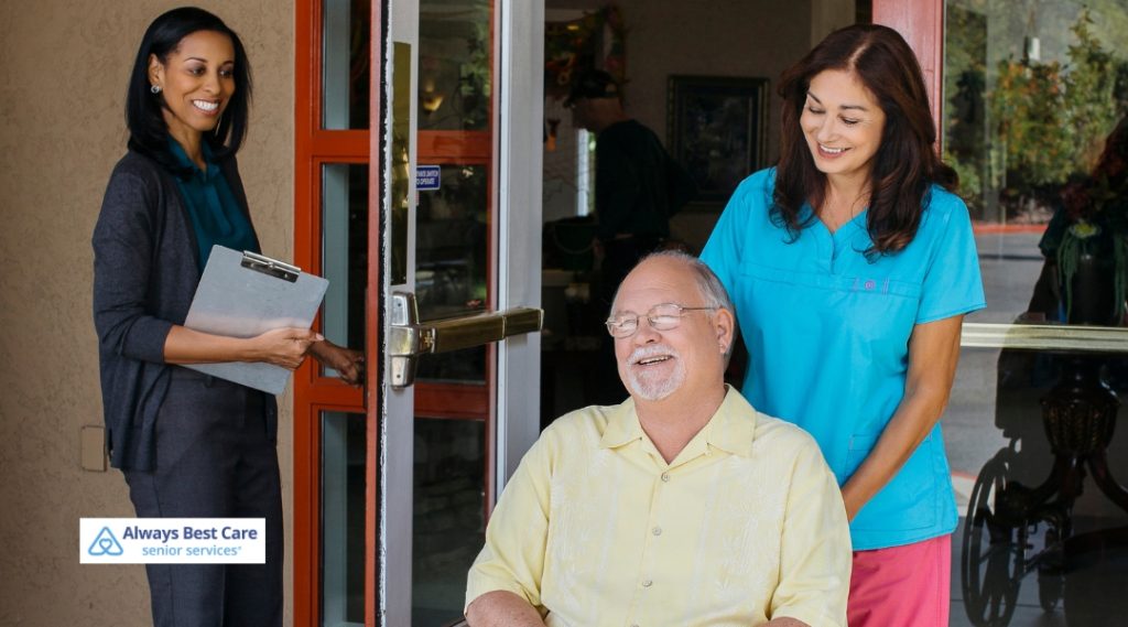 A caregiver and two family members are outside with a senior man in a wheelchair, having a conversation and sharing a joyful moment. The Always Best Care logo is in the bottom left corner.