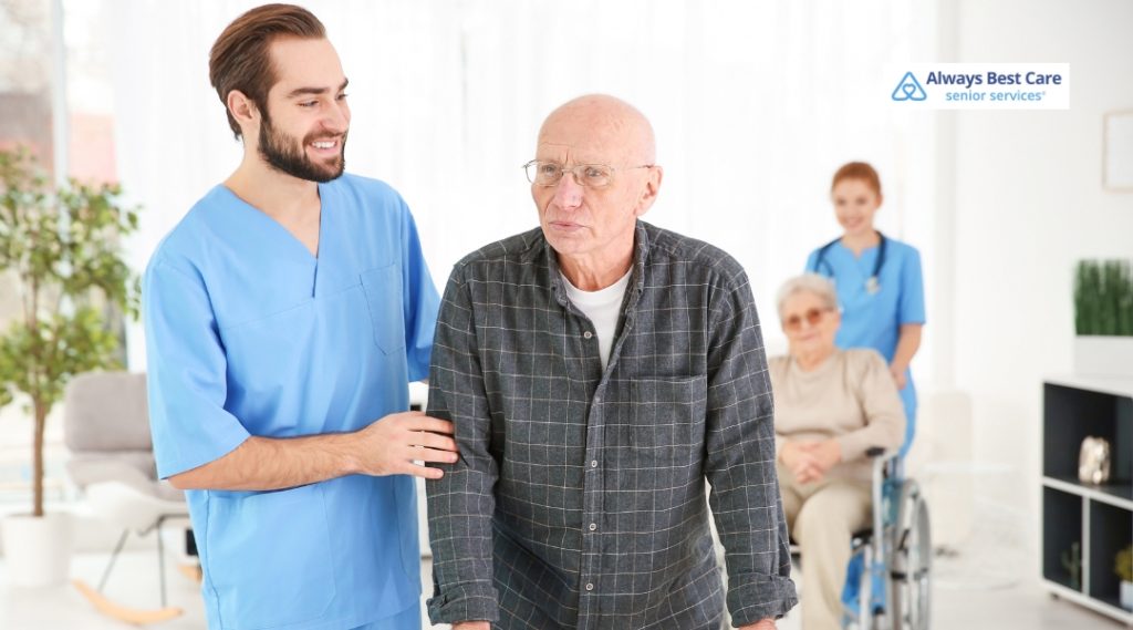 A caregiver assists an elderly man with walking, offering support and guidance as he moves through a bright and welcoming indoor space. In the background, another caregiver helps a woman in a wheelchair, reflecting the agency's comprehensive care services.