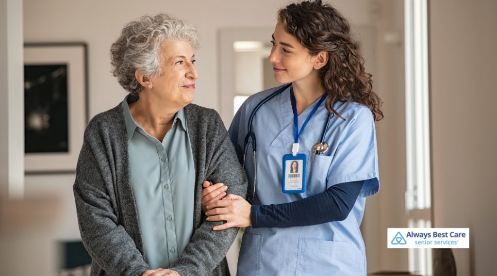 A caregiver helps an elderly woman walk with support, offering reassurance and companionship. The indoor setting is bright and calm, fostering a sense of comfort and safety for both the senior and their caregiver.