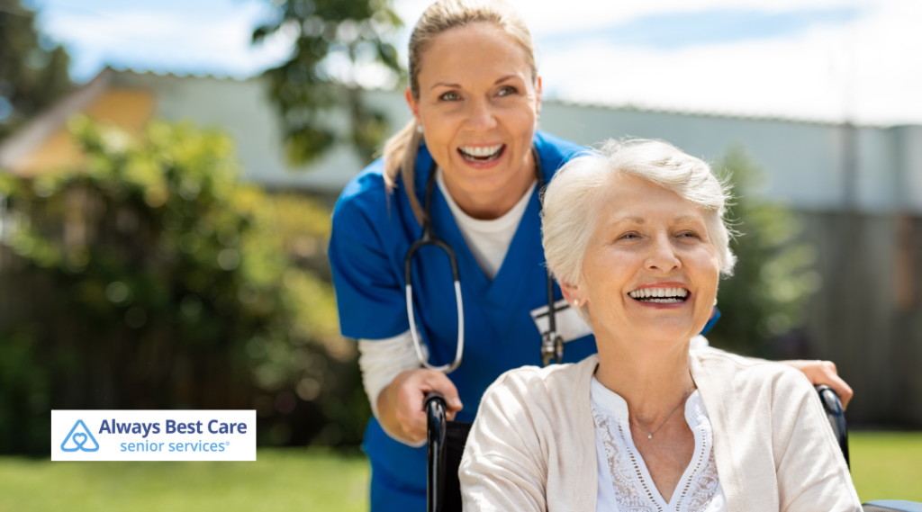 A caregiver in blue scrubs pushes a smiling elderly woman in a wheelchair outdoors, with a sunny garden and greenery in the background.