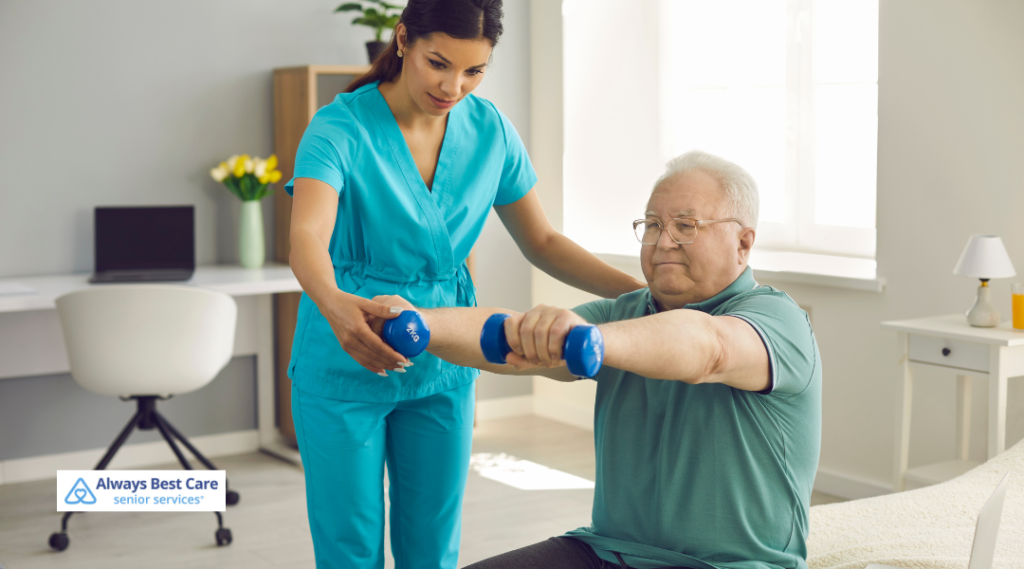 A caregiver in teal scrubs assists an elderly man with light weightlifting exercises, supporting his arm as he lifts small dumbbells in a bright, well-lit room.