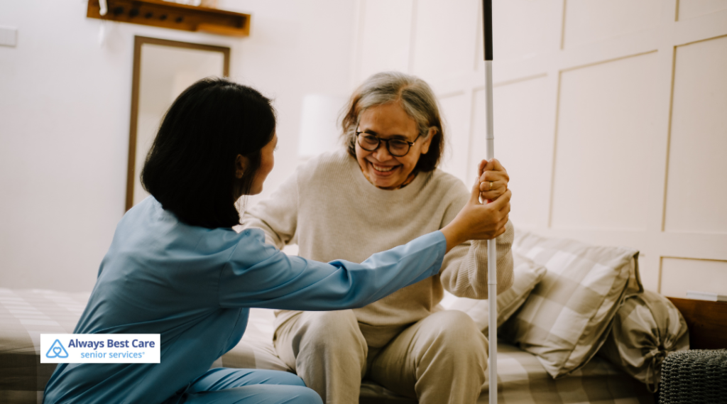 A caregiver helping a senior woman with vision support, assisting her with a walking aid in a home setting, ensuring safety and promoting independence. Always Best Care senior services logo visible at the bottom.
