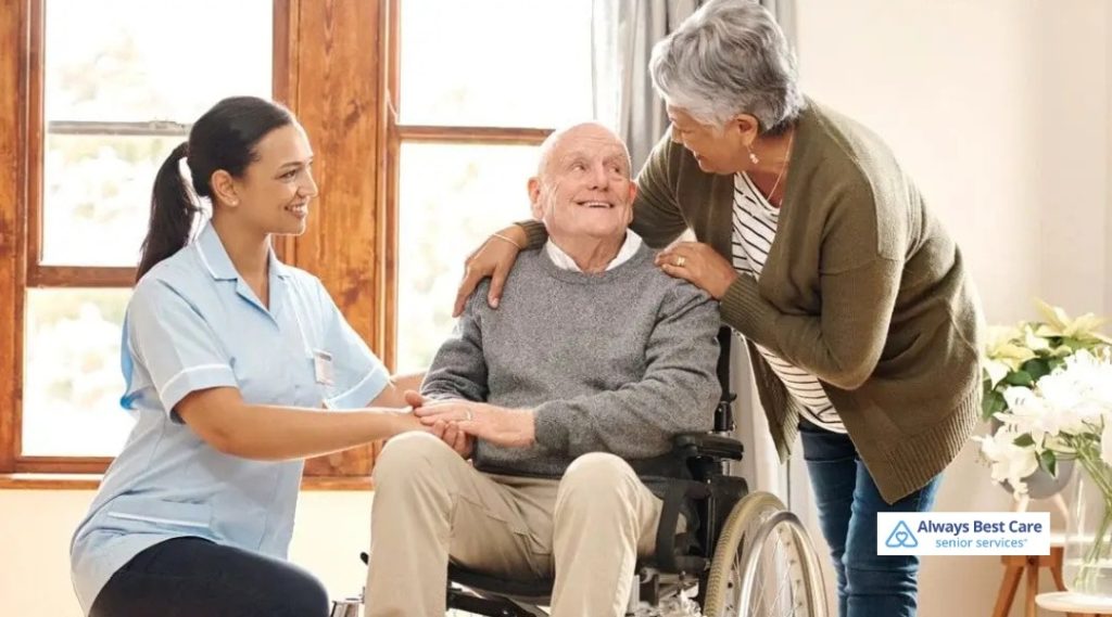 Caregiver and family member supporting an elderly man in a wheelchair at home, smiling and holding hands during a compassionate visit.