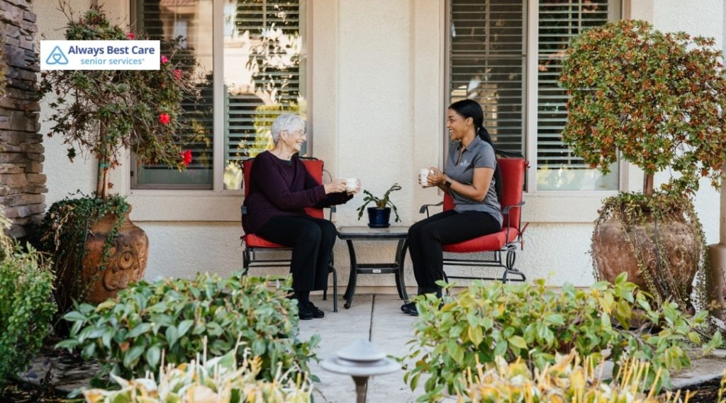 Older woman and caregiver sitting on red patio chairs, sharing hot drinks and talking in a landscaped garden outside a home with the Always Best Care Senior Services logo in the corner.