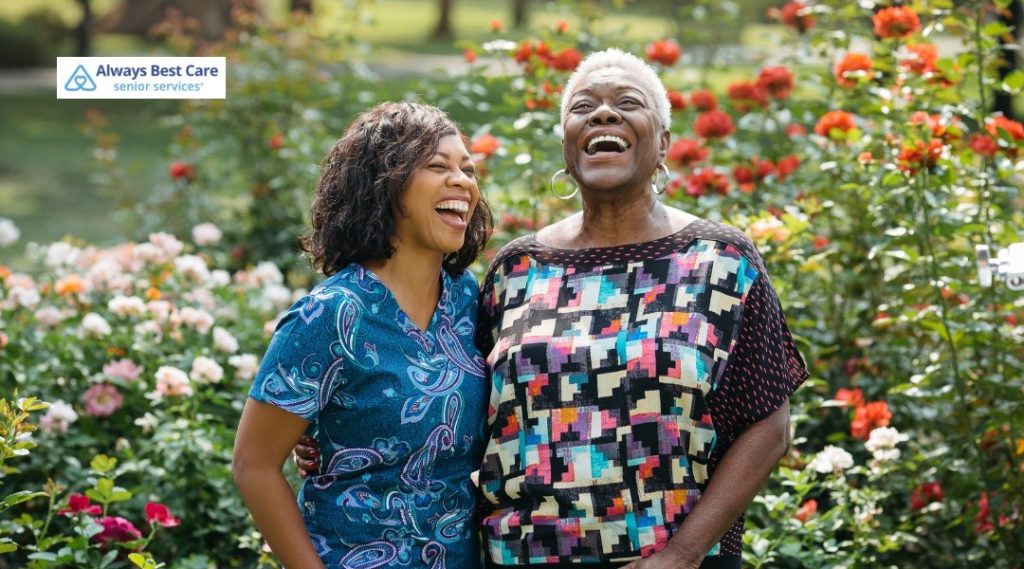 Home care professional standing in a garden beside a smiling senior woman surrounded by blooming flowers.