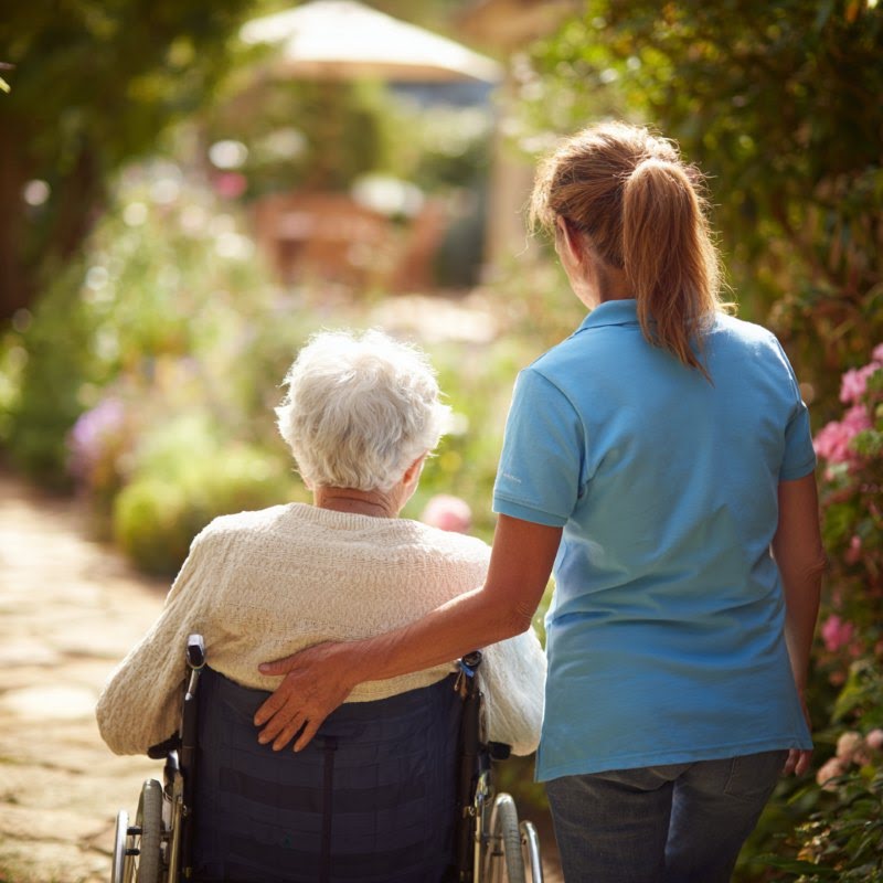 HOME CARE CHESAPEAKE, VA CAREGIVER WALKING ALONGSIDE AN ELDERLY PERSON IN A WHEELCHAIR ON A BRIGHT SUNNY OUTDOOR PATH
