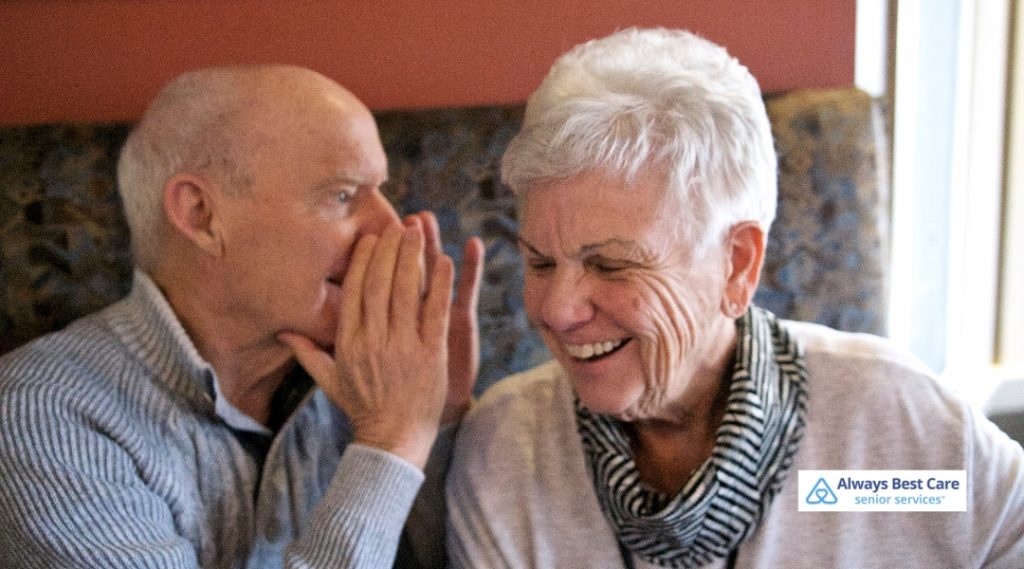 A senior man and woman share a moment of joy as the man whispers in the woman's ear, both smiling and laughing.