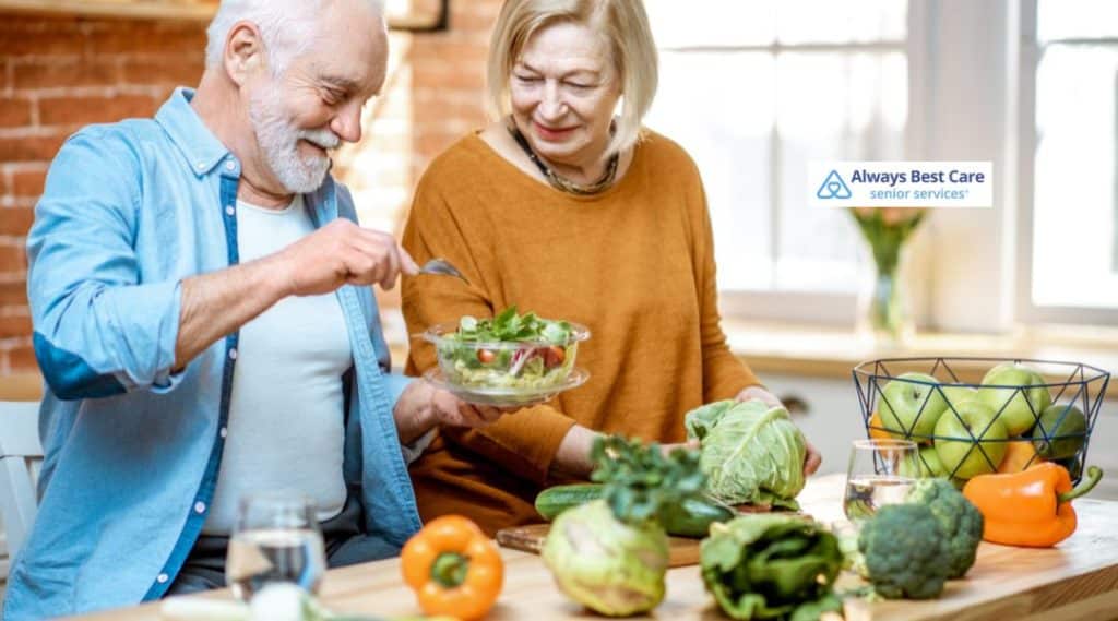 A senior couple enjoying time together in the kitchen, with the man serving a fresh salad while the woman watches and smiles. The kitchen is filled with fresh vegetables, showcasing a healthy and active lifestyle.