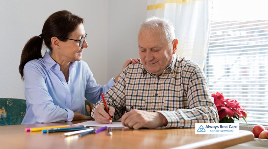 CAREGIVER SITS BESIDE AN OLDER MAN AS HE DRAWS WITH COLORED PENCILS AT A TABLE; THE ALWAYS BEST CARE SENIOR SERVICES LOGO APPEARS IN THE LOWER RIGHT.