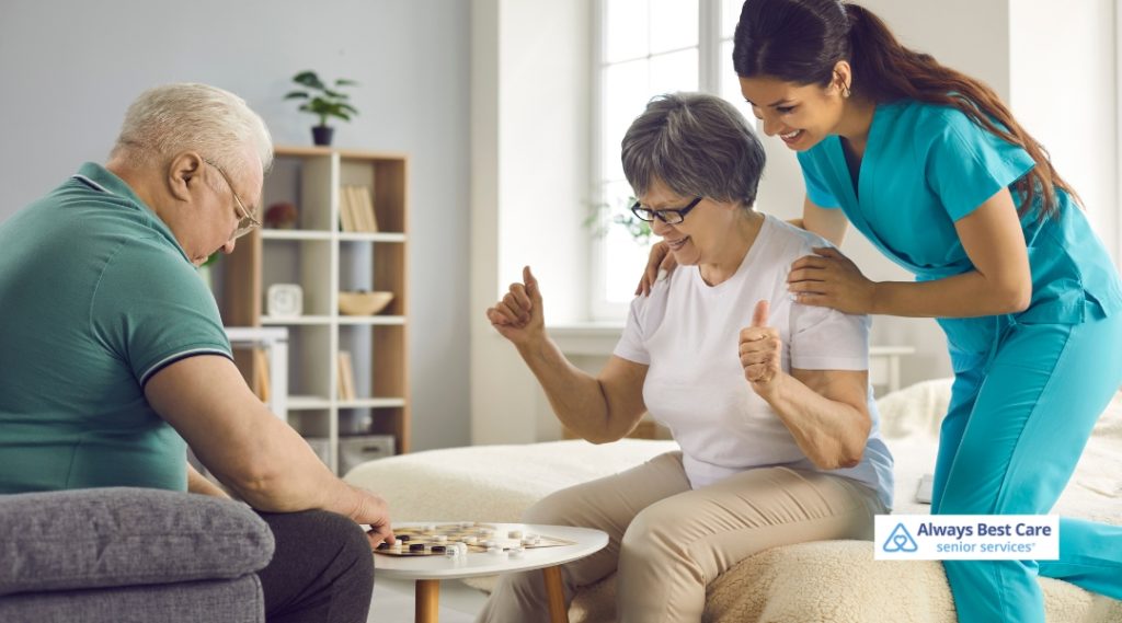A caregiver and a senior woman celebrate a game victory together, with another person playing in the background, all smiling in a bright and cheerful living room.
