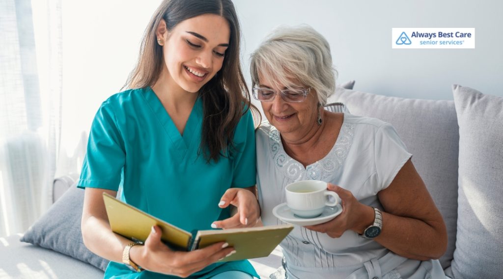 A caregiver and a senior woman are smiling and enjoying each other's company as they look at a photo album, with the senior woman holding a cup of tea.