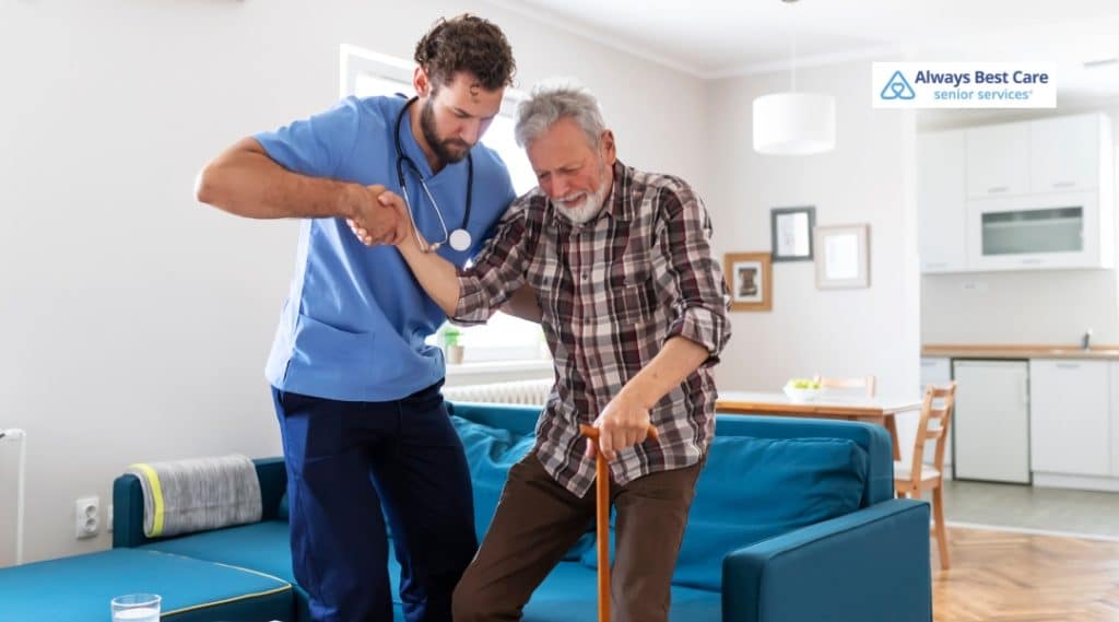 A caregiver assists an elderly man with walking inside a well-lit living room, supporting him as he uses a cane. The caregiver is wearing a blue medical uniform and offering steady support with a friendly expression.