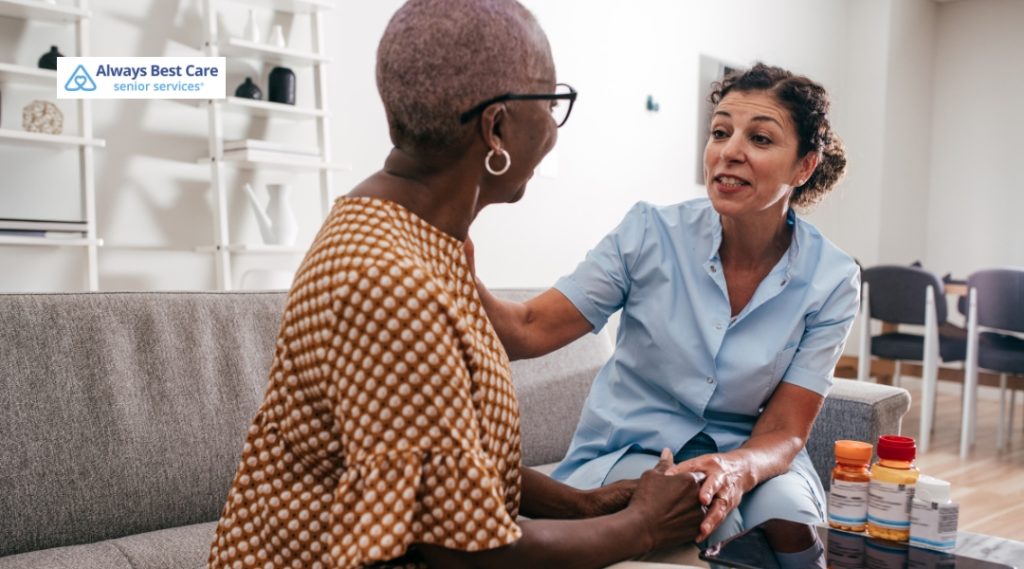 A caregiver is sitting with a senior woman, helping her with medication management. The caregiver, in a light blue uniform, is smiling and engaging in conversation, while the elderly woman looks attentively, with several medication bottles on the table.