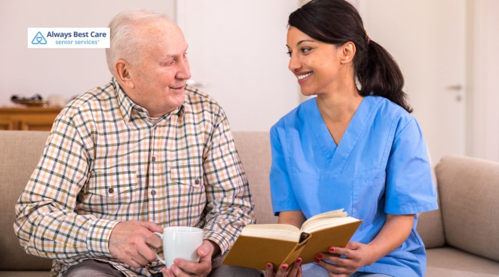 A caregiver and an elderly man share a cheerful moment outdoors. The caregiver, in a blue uniform, supports the man, who is smiling and looking happy, as they walk together through a lush garden.