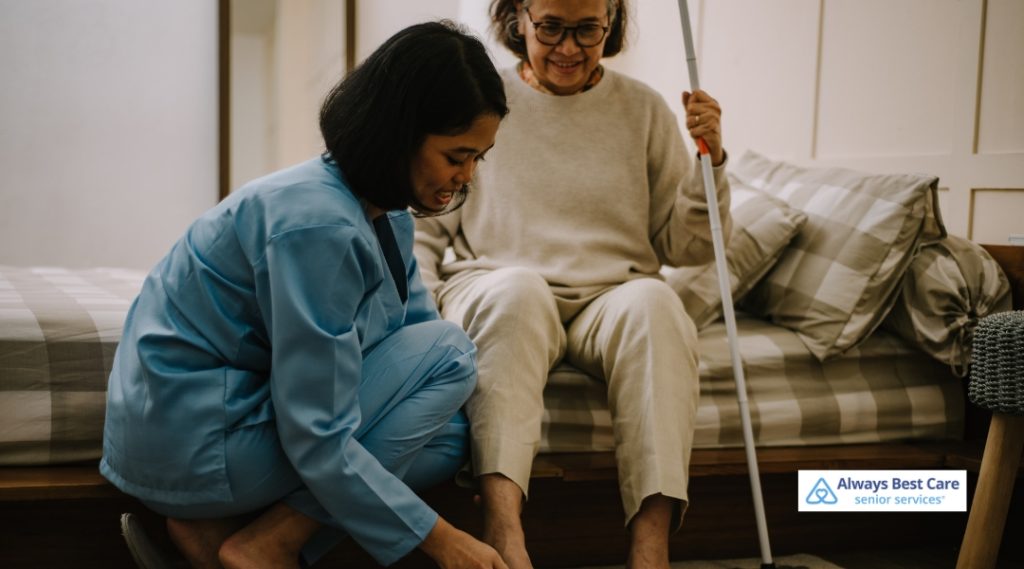 A caregiver helps a senior woman with her footwear while sitting on a bed in a cozy bedroom. The woman looks comfortable in her light-colored clothes, and the caregiver, dressed in a blue uniform, is focused on assisting her.