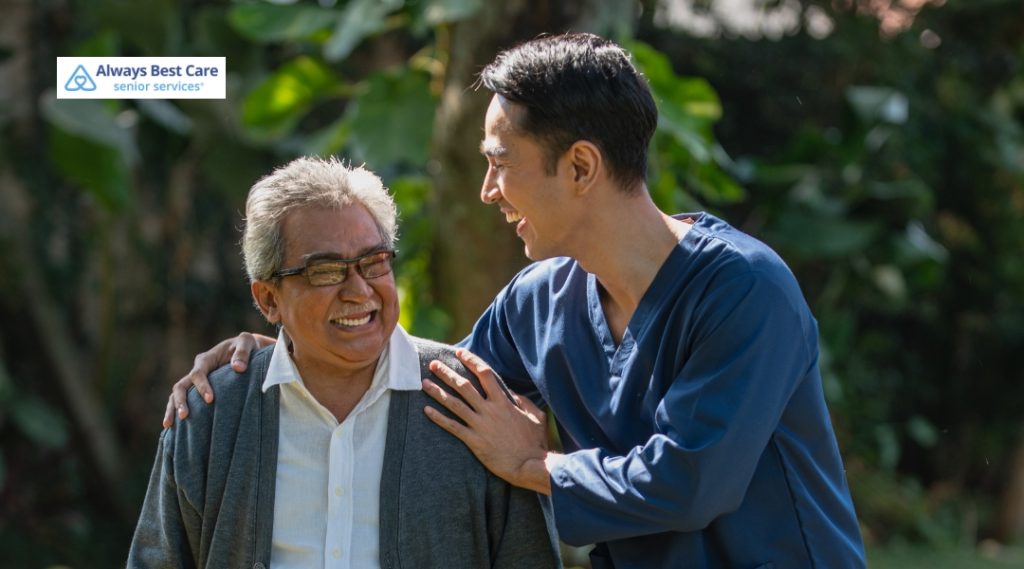 A caregiver and an elderly man share a cheerful moment outdoors. The caregiver, in a blue uniform, supports the man, who is smiling and looking happy, as they walk together through a lush garden.
