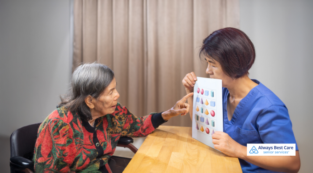 Always Best Care nurse in blue scrubs holding a sheet of shapes while an elderly woman with dementia points to images during a simple memory and recognition exercise.”