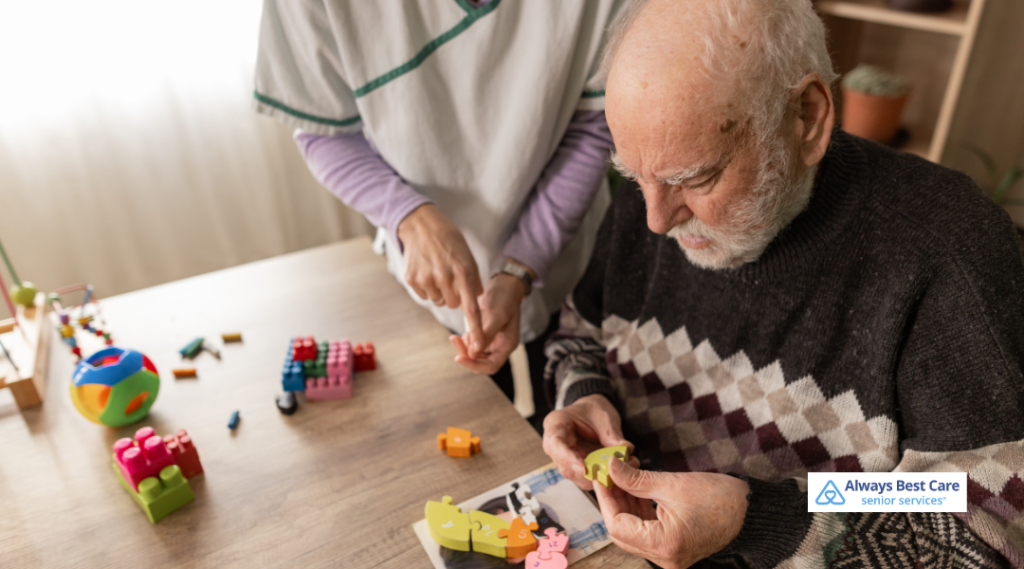Senior man with memory loss working on colorful puzzle pieces at a table while an Always Best Care dementia caregiver offers gentle guidance beside him.”