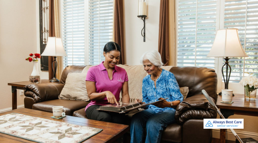 Always Best Care caregiver in pink polo sitting on a couch with an older woman, looking through a photo album together to support memory and reminiscence.
