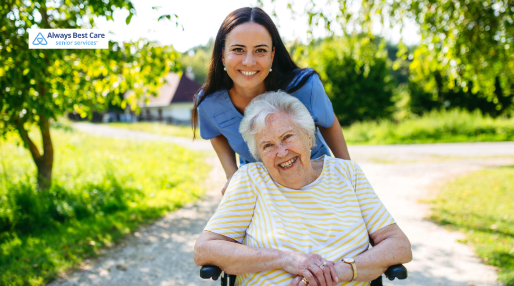 Caregiver in blue scrubs behind elderly woman in a wheelchair smiling in a park
