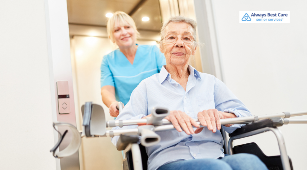 Caregiver in teal scrubs wheels a smiling senior woman out of an elevator, with her crutches resting across the wheelchair.