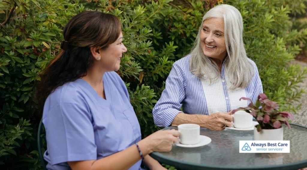 Caregiver and senior woman sitting outside at a patio table, enjoying coffee together in a lush garden setting, Always Best Care logo in corner.