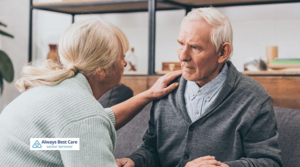 Older woman gently touching an older man’s shoulder, offering comfort as they sit together on a couch