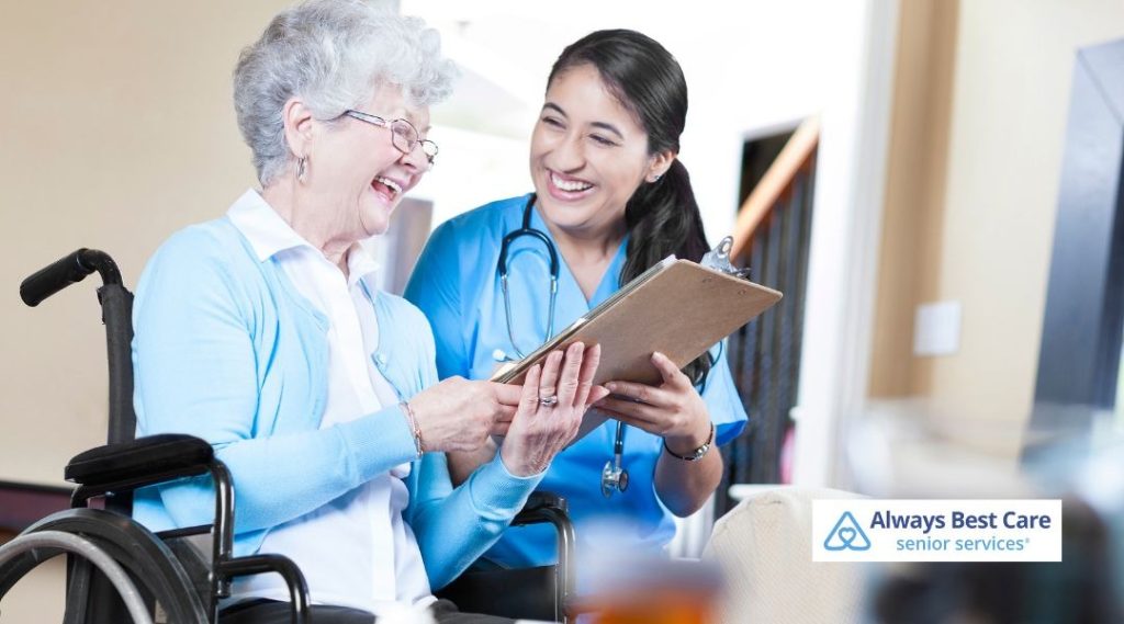 Older woman in a wheelchair laughing with a nurse who is holding a clipboard as they share a lighthearted moment.