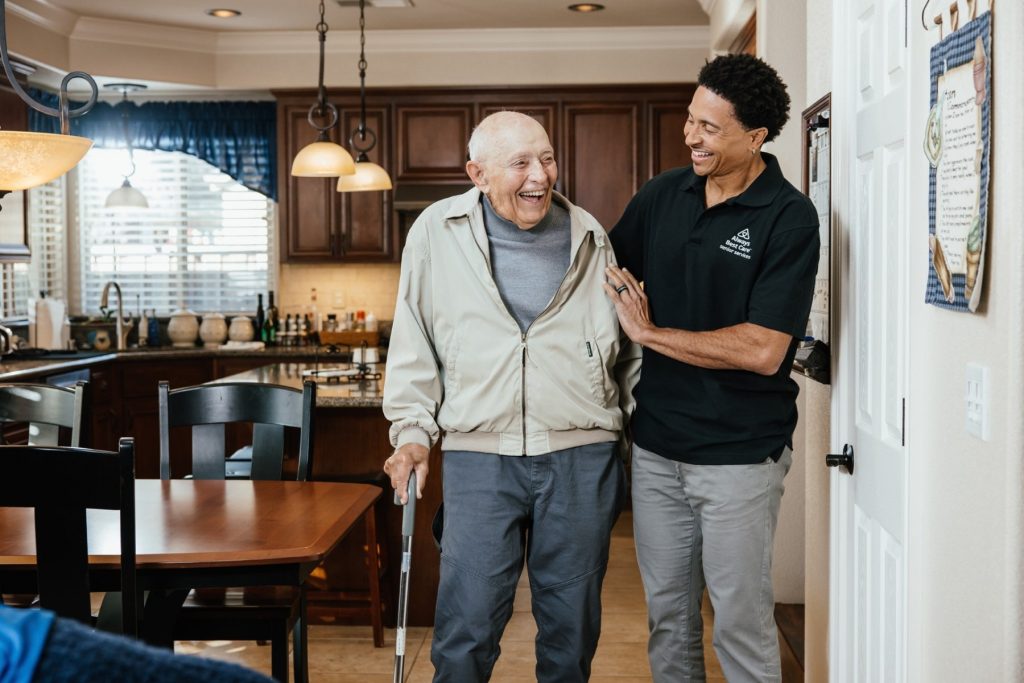 Caregiver walks beside an older man using a cane in his home kitchen.