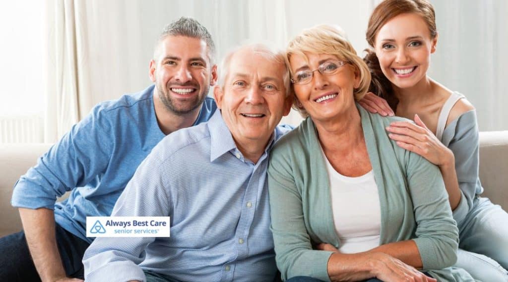 A smiling family of four, including an older couple and two younger adults, enjoying time together on a couch. The image features the Always Best Care logo at the bottom-left corner, emphasizing the connection between family and senior care.