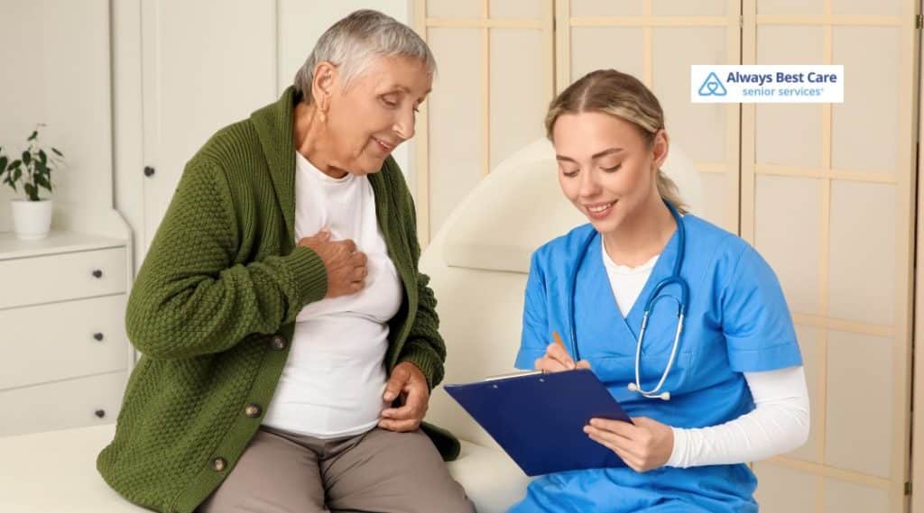 A nurse in scrubs assisting an elderly woman who is sitting on an examination table, with the nurse taking notes. The image reflects compassionate care and the importance of medical attention for seniors, with the Always Best Care logo visible.