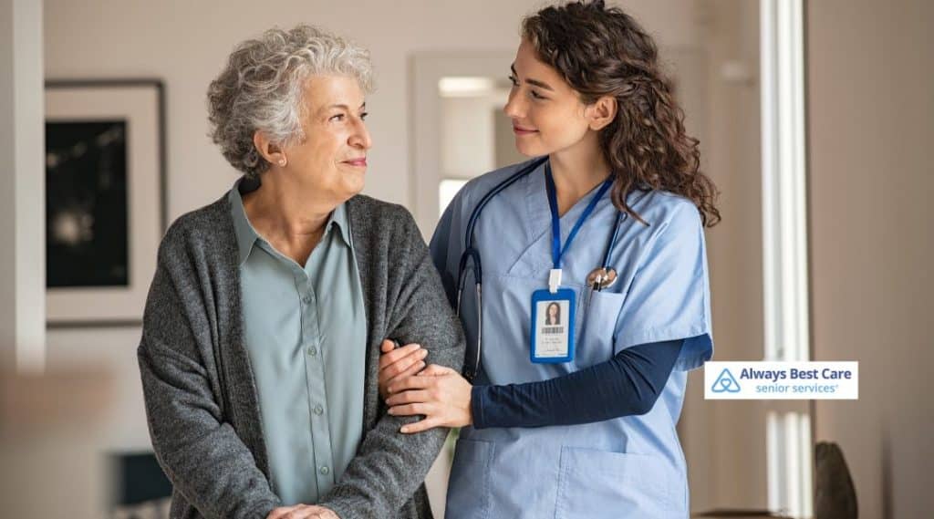A caregiver is gently assisting a senior woman in walking, offering support as the senior woman smiles. The image highlights the trust and support provided by Always Best Care, with the logo in the bottom-right corner.