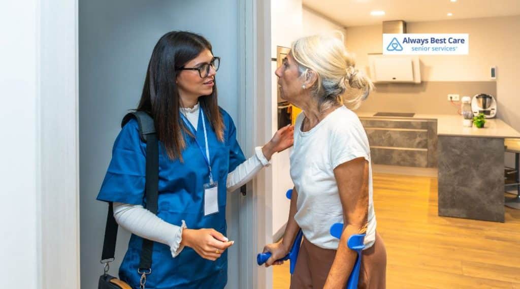 A caregiver in scrubs speaks with a senior woman holding crutches, providing assistance and offering a warm interaction. The Always Best Care logo is placed at the top-right corner, signaling the importance of compassionate home care.