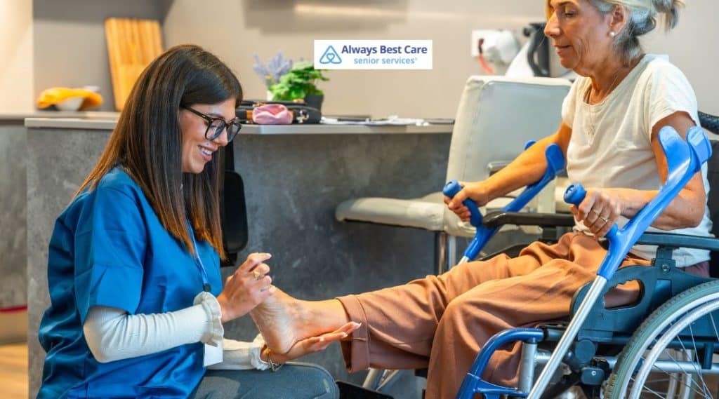 A caregiver gently cares for the foot of an elderly woman sitting in a wheelchair, highlighting personalized care and attention to detail. The Always Best Care logo is located at the top-left corner, reinforcing the brand’s commitment to senior well-being.