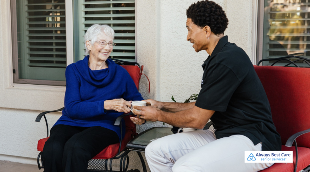 Always Best Care caregiver offering a warm drink to a smiling senior during an outdoor companion care moment.