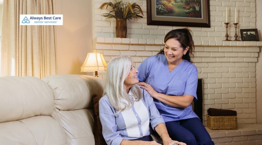 A senior woman and her caregiver, smiling and engaging in a pleasant conversation while sitting on a couch in a cozy, well-decorated living room. The Always Best Care logo is in the top left corner.
