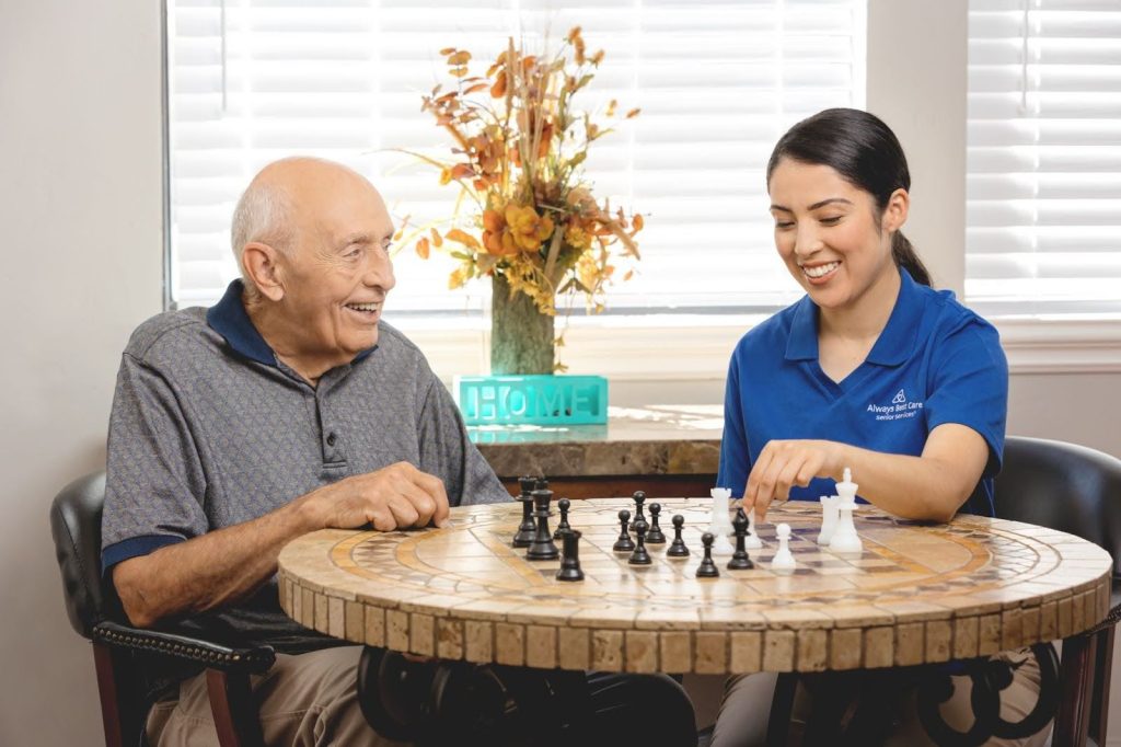 Caregiver playing a game of chess with elderly man, both smiling and engaging in the activity, with the Always Best Care logo visible.