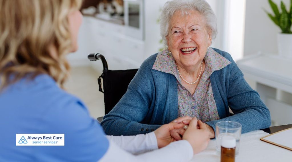 Older woman smiling warmly while holding hands with a caregiver during a friendly conversation at a table.
