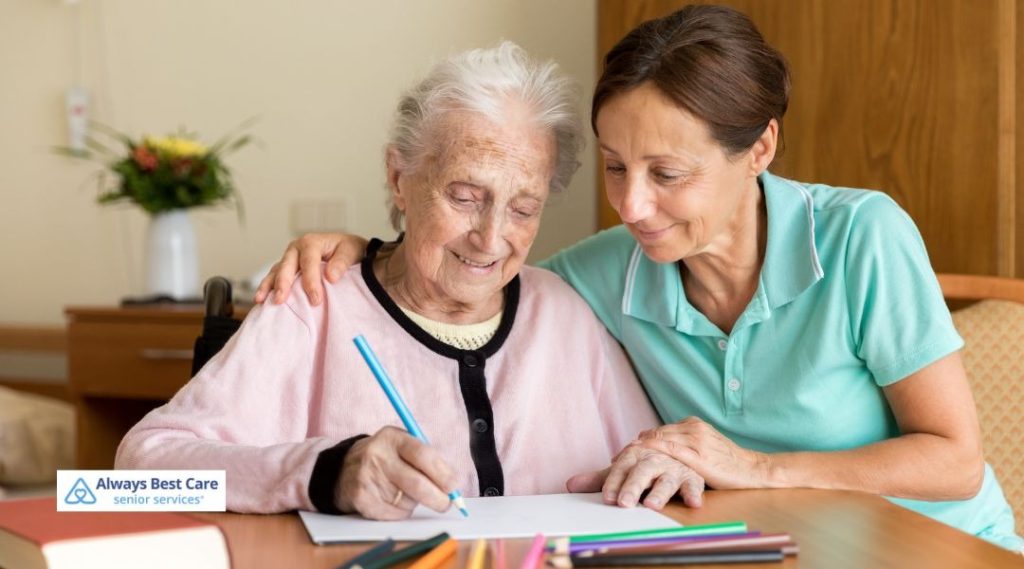 Caregiver supporting an older woman during a creative activity, helping her draw with colored pencils at a table.