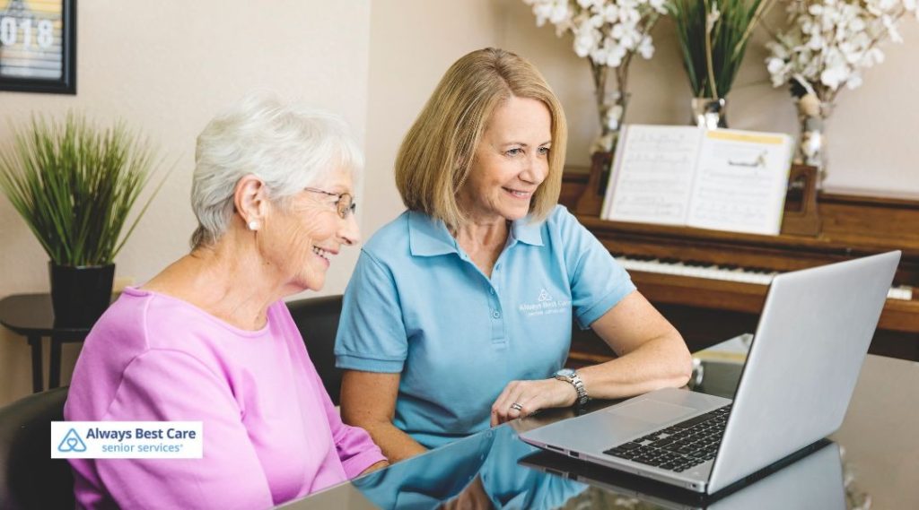 Caregiver sitting beside an older woman, helping her use a laptop at home while offering guidance and companionship.