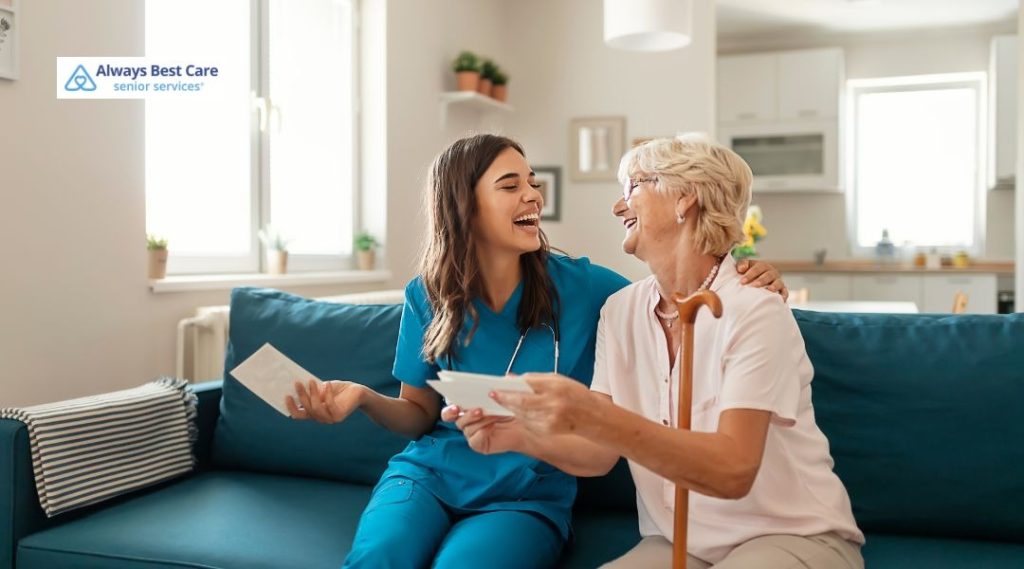 Female caregiver in blue scrubs sitting on a couch with a senior woman using a cane as they look at papers or photos together in a bright living room, Always Best Care logo in the corner.