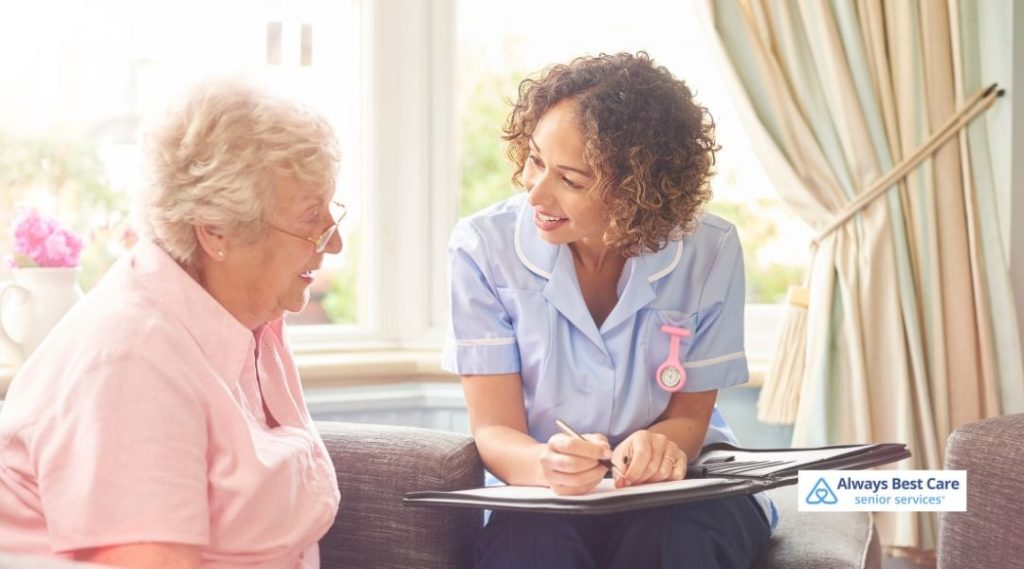 Senior woman sitting on a sofa while a female caregiver in a light blue uniform takes notes on a clipboard during a home care consultation, Always Best Care logo in the corner.