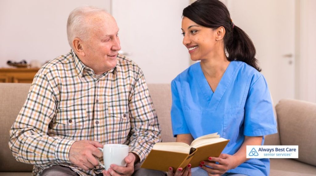 Female caregiver in blue scrubs sitting closely with a smiling senior woman with a cane as they look at papers together on a teal sofa in a cozy home, Always Best Care logo in the corner.