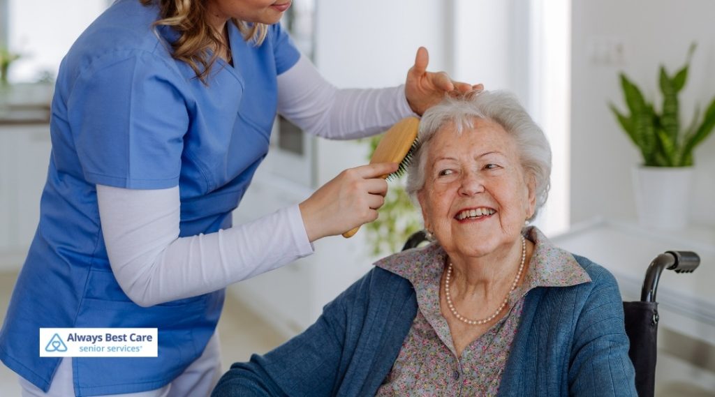 Caregiver brushing an older woman’s hair while she sits in a wheelchair in a bright home, providing gentle personal care, Always Best Care logo in corner.