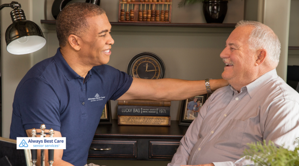 A friendly caregiver sits beside an older man, both smiling and engaged in a relaxed conversation inside a cozy living room filled with books and warm lighting. The Always Best Care logo is shown in the lower left.