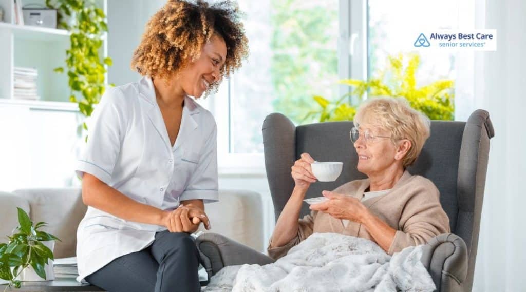 Caregiver chats with an older woman sipping tea in a cozy chair, offering companionship and support.