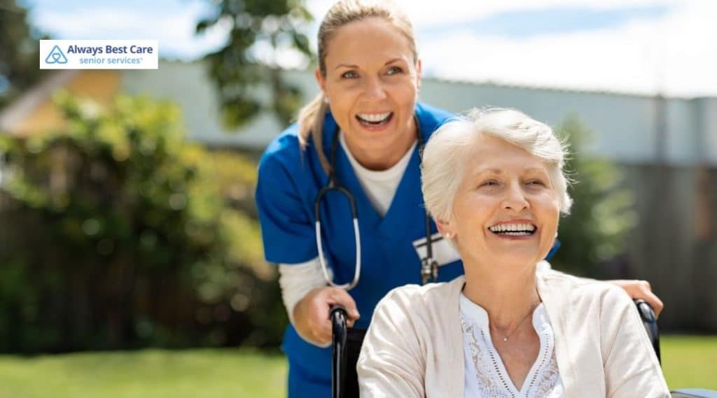 Nurse pushes a smiling older woman in a wheelchair outdoors on a sunny day.