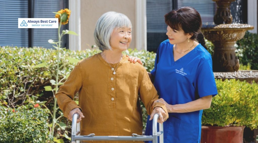 emale caregiver in blue medical scrubs gently supporting an older woman using a walker in a sunny garden courtyard, with the Always Best Care Senior Services logo in the corner.