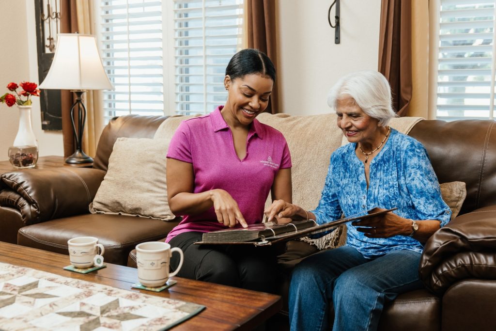 A caregiver in a pink polo sits with an older woman on a sofa, looking through a photo album together with coffee cups on the table.