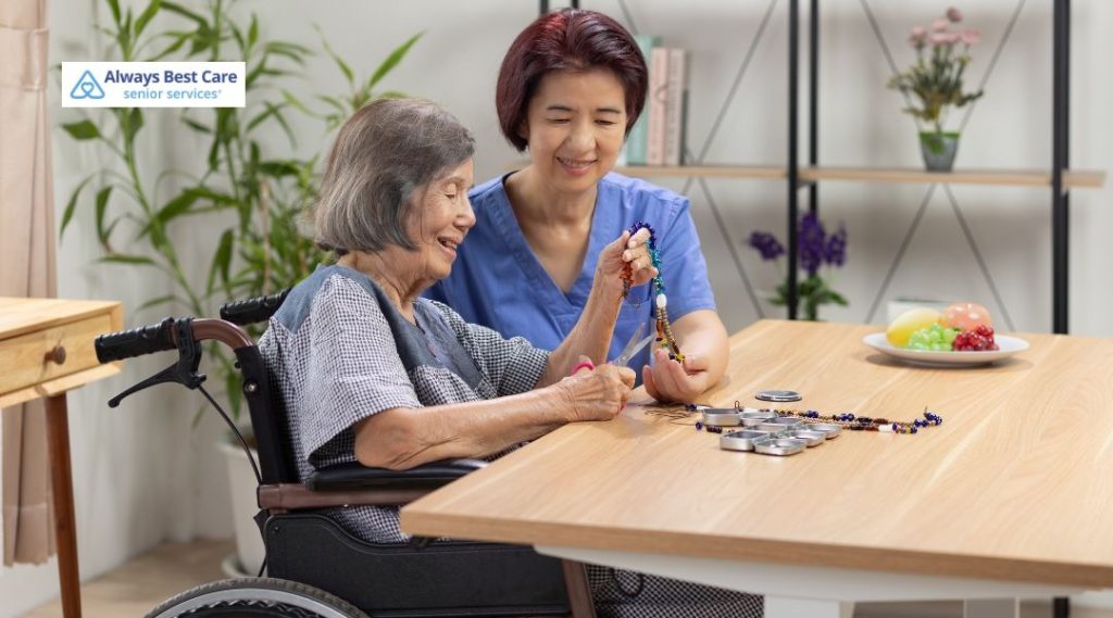 A caregiver in blue scrubs helps an older woman in a wheelchair with a colorful beading craft project at a table.